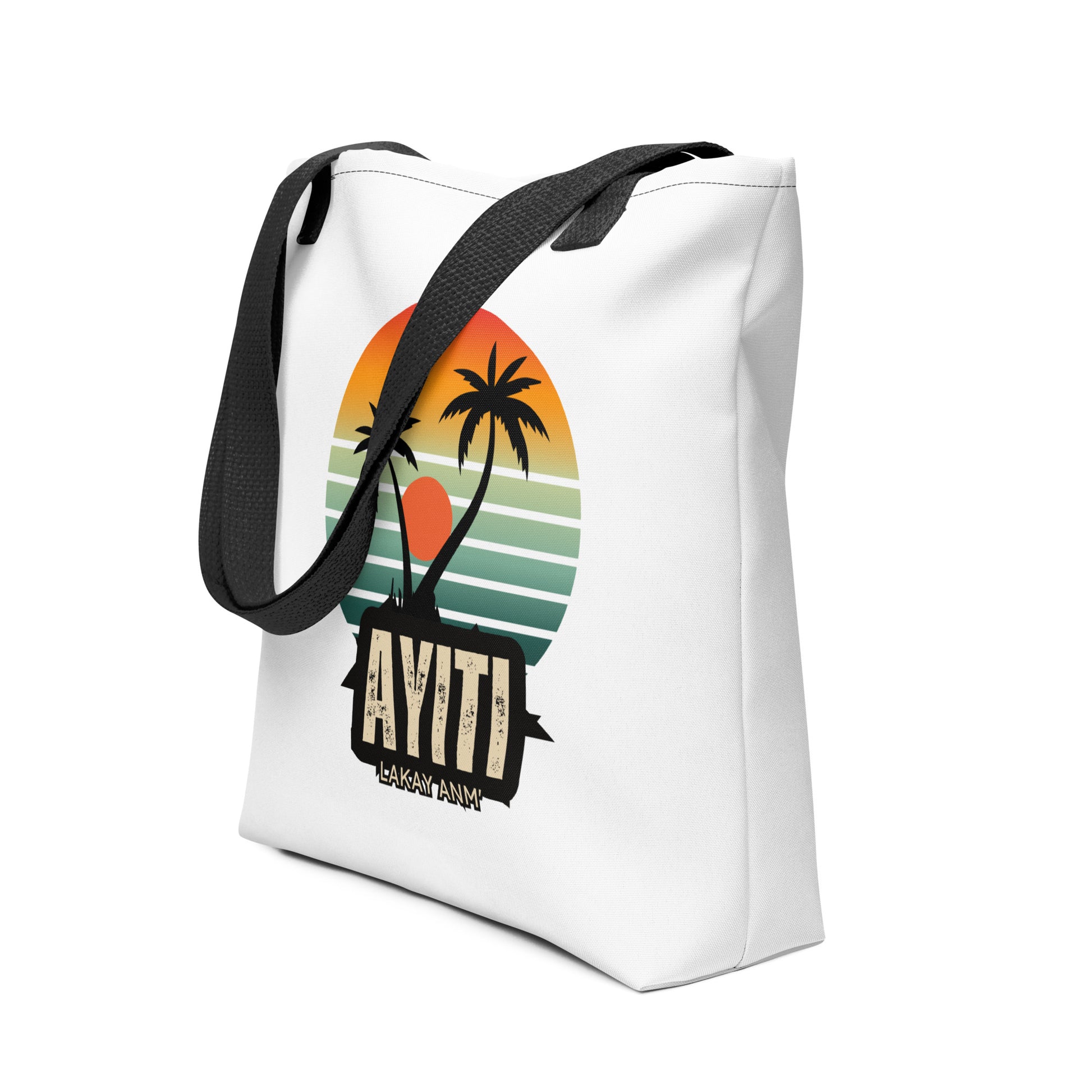 White canvas tote bag with black handles (left side view), featuring a circular graphic with an orange and yellow sunset over blue and green stripes, silhouetted by two palm trees. The text 'AYITI' and 'LAKAY ANM'' is below the graphic in a stylized font.