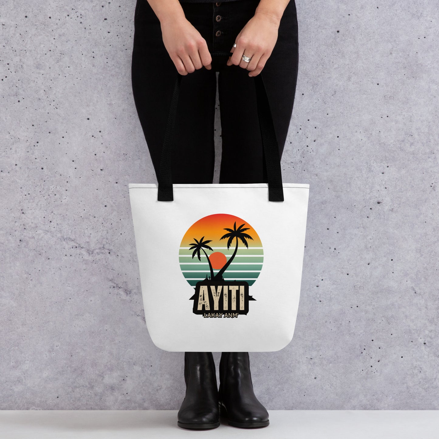 Model holding a White canvas tote bag with black handles, featuring a circular graphic with an orange and yellow sunset over blue and green stripes, silhouetted by two palm trees. The text 'AYITI' and 'LAKAY ANM'' is below the graphic in a stylized font.Model is standing wearing black pants with black shoes. She's holding the bag right over her shoes