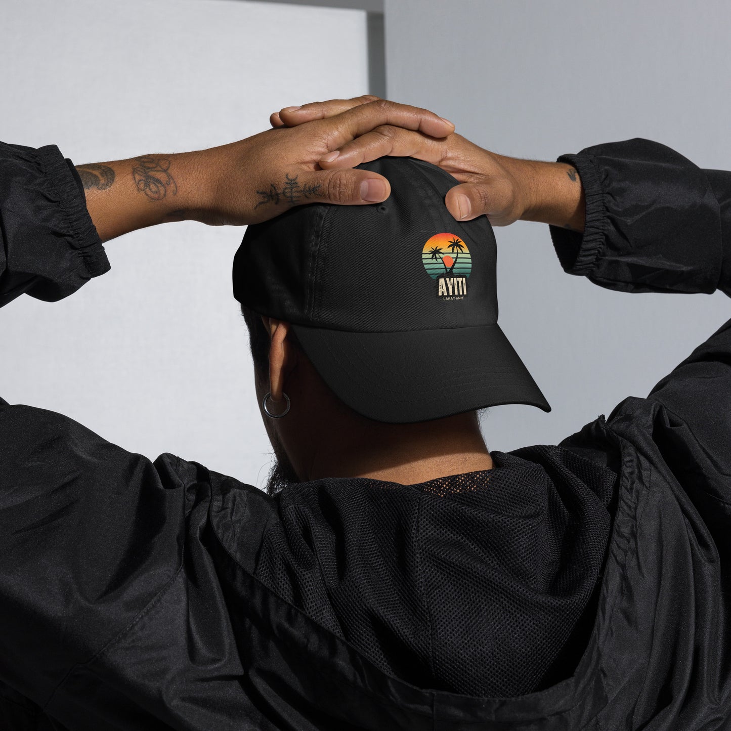 Close-up of a person wearing a black baseball cap (backwards)with a circular graphic on the front featuring an orange and yellow sunset over blue and green stripes, silhouetted by two palm trees. The text 'AYITI' and 'LAKAY ANM'' is below the graphic in a stylized font. The person is standing with their hands over their hand