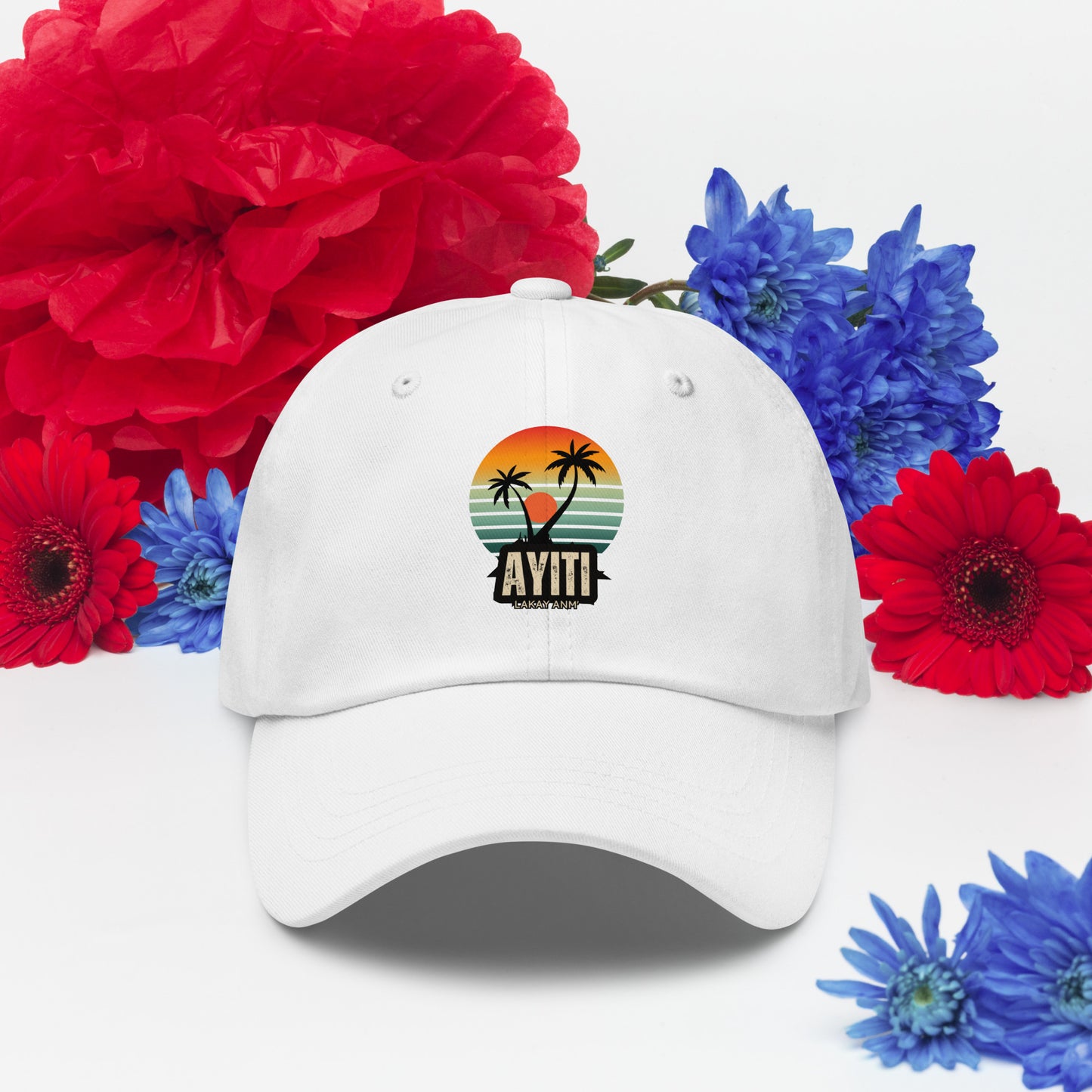 White baseball cap with a circular graphic on the front featuring an orange and yellow sunset over blue and green stripes, silhouetted by two palm trees. The text 'AYITI' and 'LAKAY ANM'' is below the graphic in a stylized font.