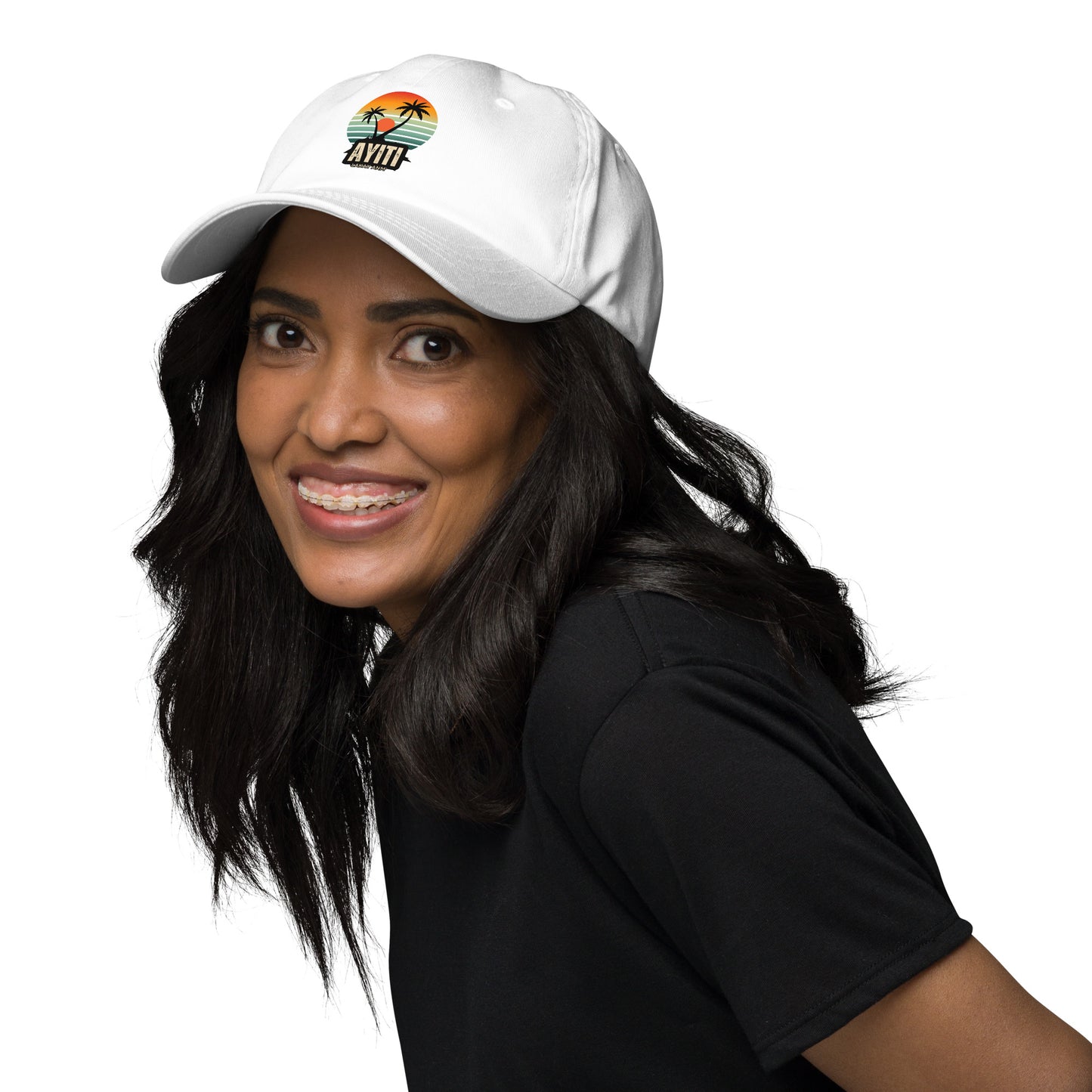 Smiling female modelwearing a white baseball cap with a circular graphic on the front featuring an orange and yellow sunset over blue and green stripes, silhouetted by two palm trees. The text 'AYITI' and 'LAKAY ANM'' is below the graphic in a stylized font.