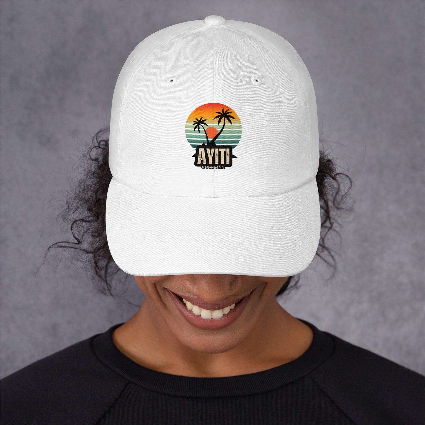 Close-up of a person wearing a white baseball cap with a circular graphic on the front featuring an orange and yellow sunset over blue and green stripes, silhouetted by two palm trees. The text 'AYITI' and 'LAKAY ANM'' is below the graphic in a stylized font. The person is smiling slightly.