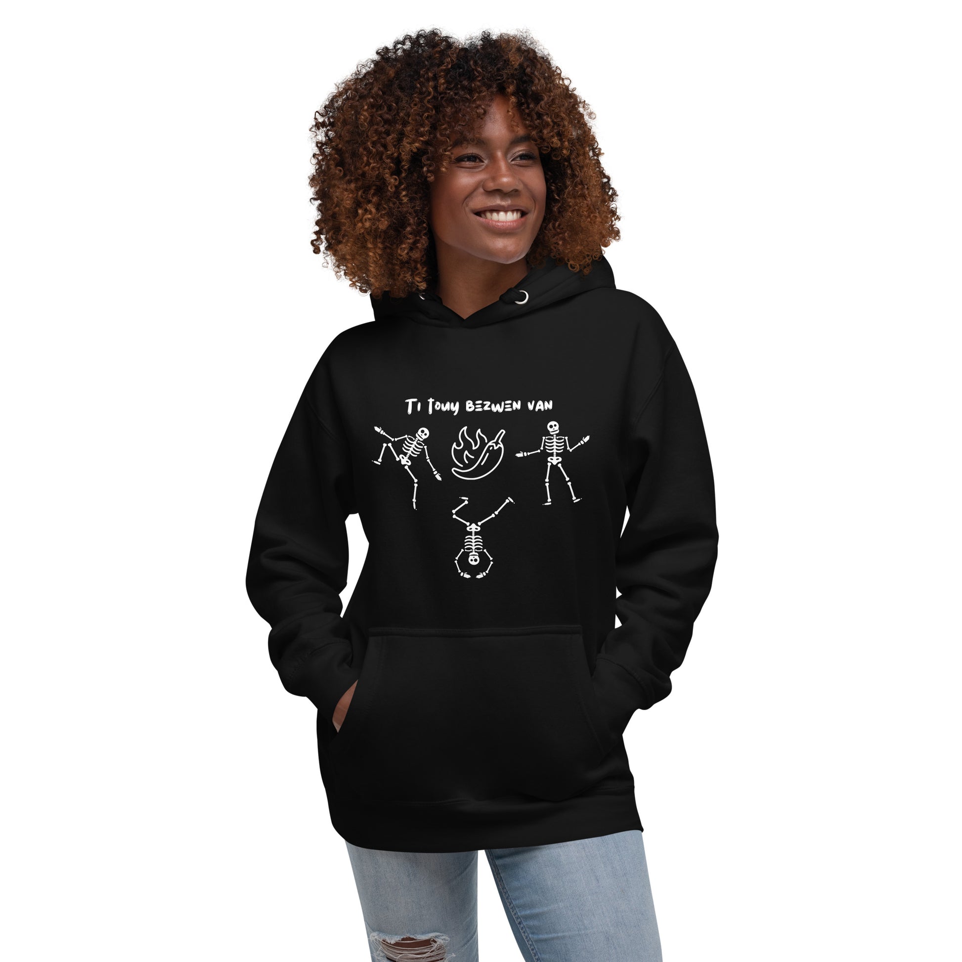 A smiling black female model wearing a pullover hoodie with a white line-art graphic of three dancing skeletons. The middle skeleton is doing a handstand. Above them is a hot pepper with flames, and the text 'TI TOUY BEZWEN NAN'. The design is a playful nod to a Haitian cultural tradition.