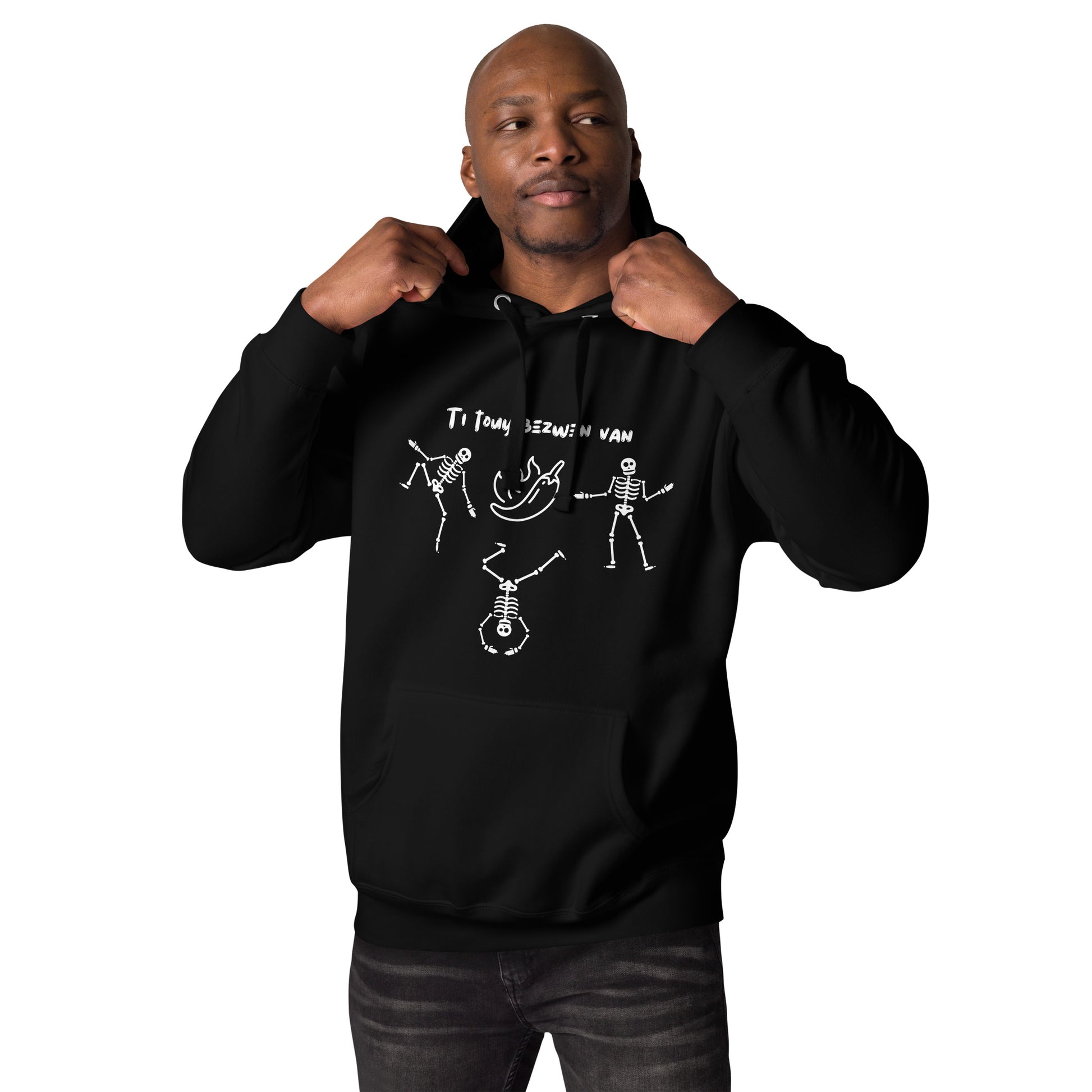 A black male model wearing a pullover hoodie with a white line-art graphic of three dancing skeletons. The middle skeleton is doing a handstand. Above them is a hot pepper with flames, and the text 'TI TOUY BEZWEN NAN'. The design is a playful nod to a Haitian cultural tradition.
