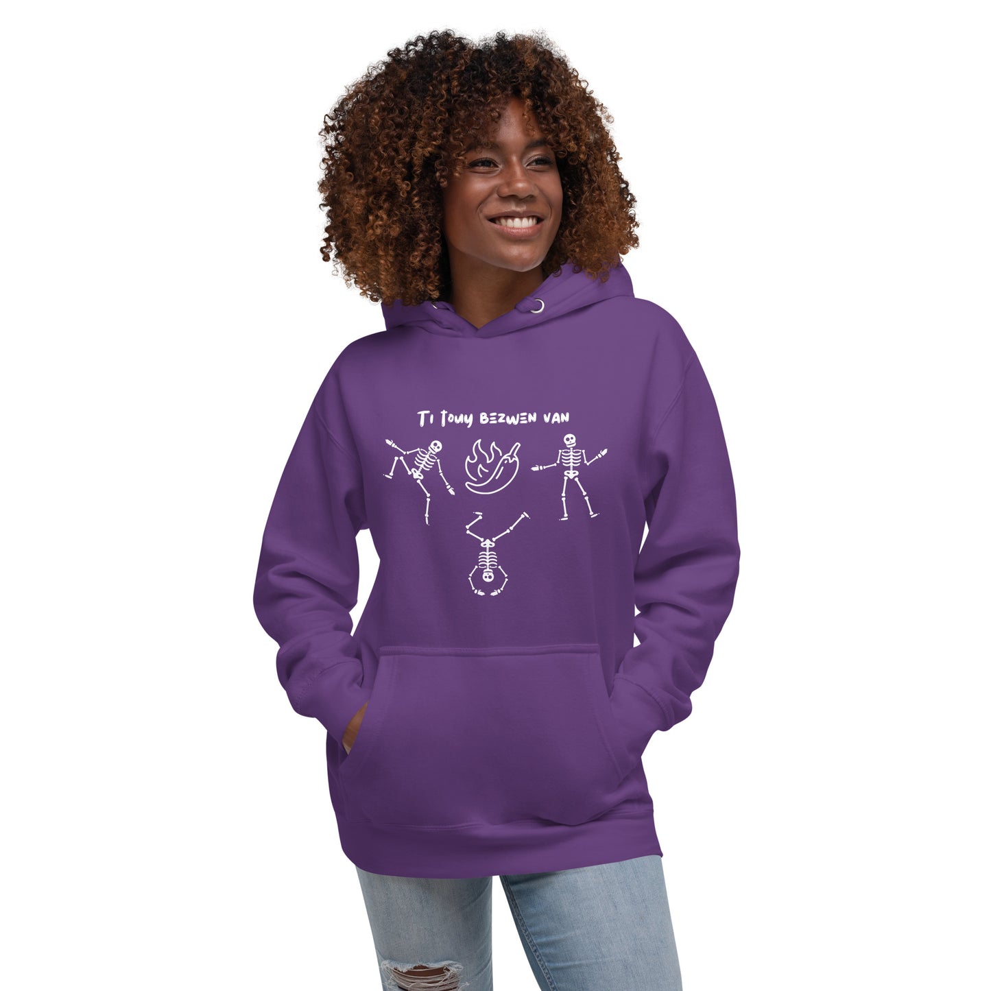 A smiling black female model wearing 3 pullover hoodie with a white line-art graphic of three dancing skeletons. The middle skeleton is doing a handstand. Above them is a hot pepper with flames, and the text 'TI TOUY BEZWEN NAN'. The design is a playful nod to a Haitian cultural tradition.