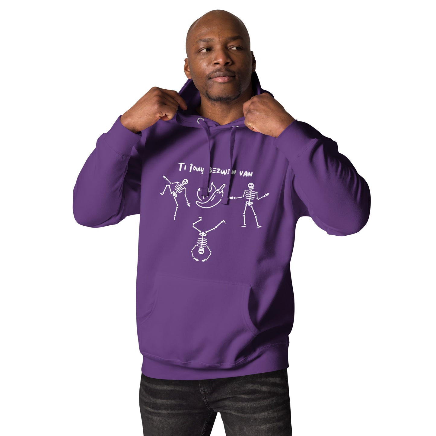 A black male model wearing a pullover hoodie with a white line-art graphic of three dancing skeletons. The middle skeleton is doing a handstand. Above them is a hot pepper with flames, and the text 'TI TOUY BEZWEN NAN'. The design is a playful nod to a Haitian cultural tradition.