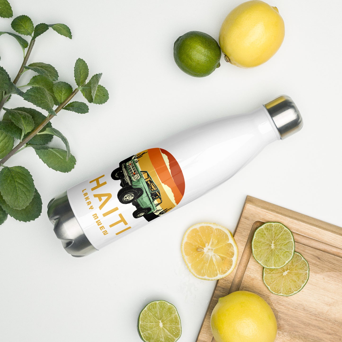 A white insulated water bottle with a silver cap and base, featuring a circular orange and yellow sunset graphic with a green vintage Jeep, black mountains, and cactus silhouettes. The text 'HAITI' is below the graphic in orange, with 'LAKAY MWEN' underneath it. A mint bunch, a lime and lime slices, and two lemons and a lemon slice are on the surface next to the bottle.