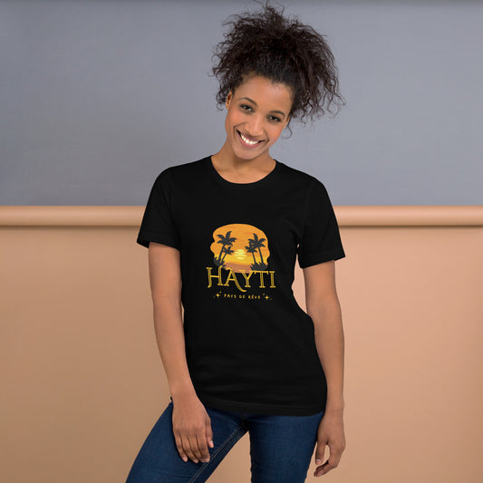 Smiling woman wearing a black Bella+Canvas 'HAYTI PAYS DE RÊVE' unisex T-shirt (front view) featuring a golden hour sunset graphic with palm trees and twinkling stars, in a room with a Grey and beige background.