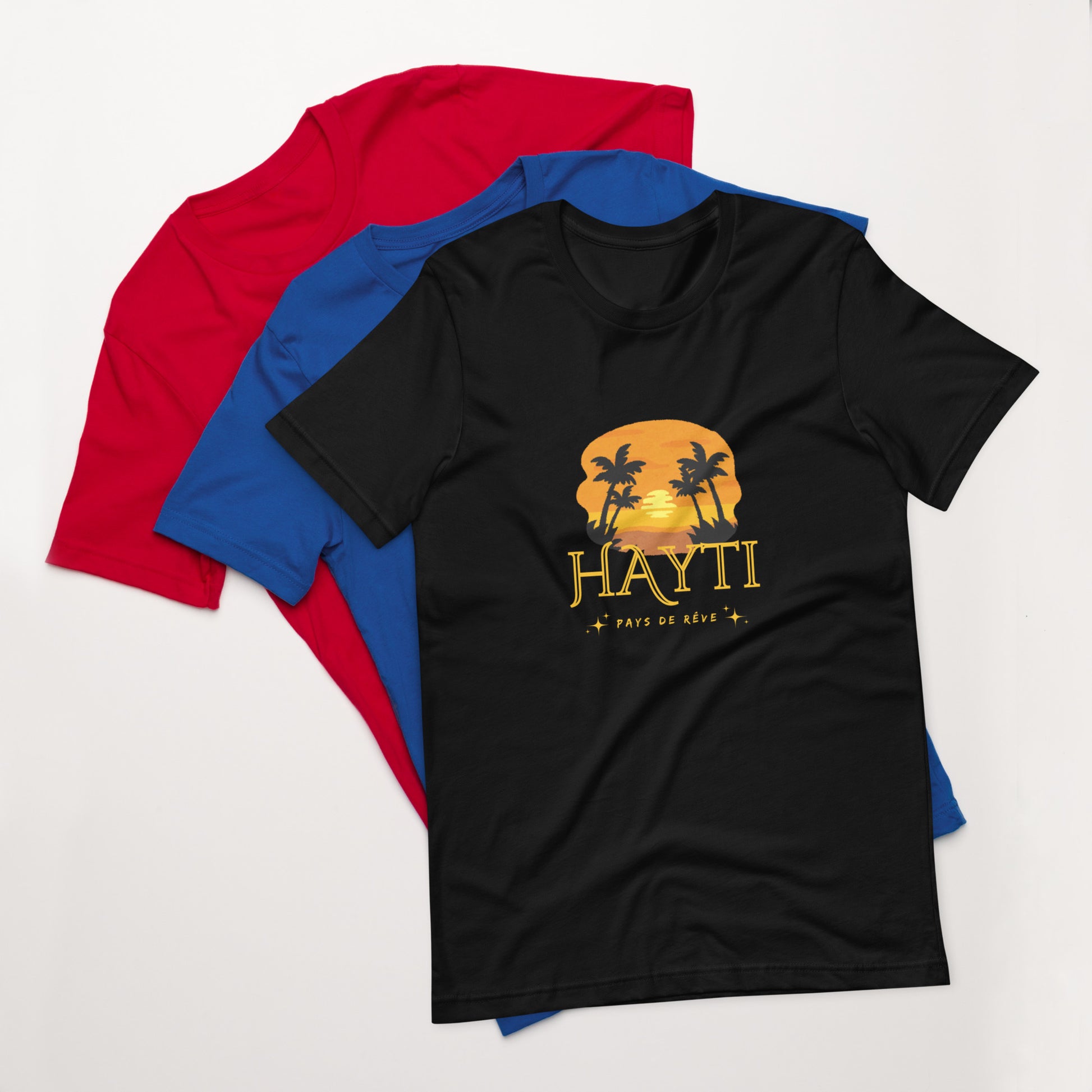 A black Bella+Canvas 'HAYTI PAYS DE RÊVE' unisex T-shirt (front view) featuring a golden hour sunset graphic with palm trees and twinkling stars. Red and bleu T-shirt in the background.