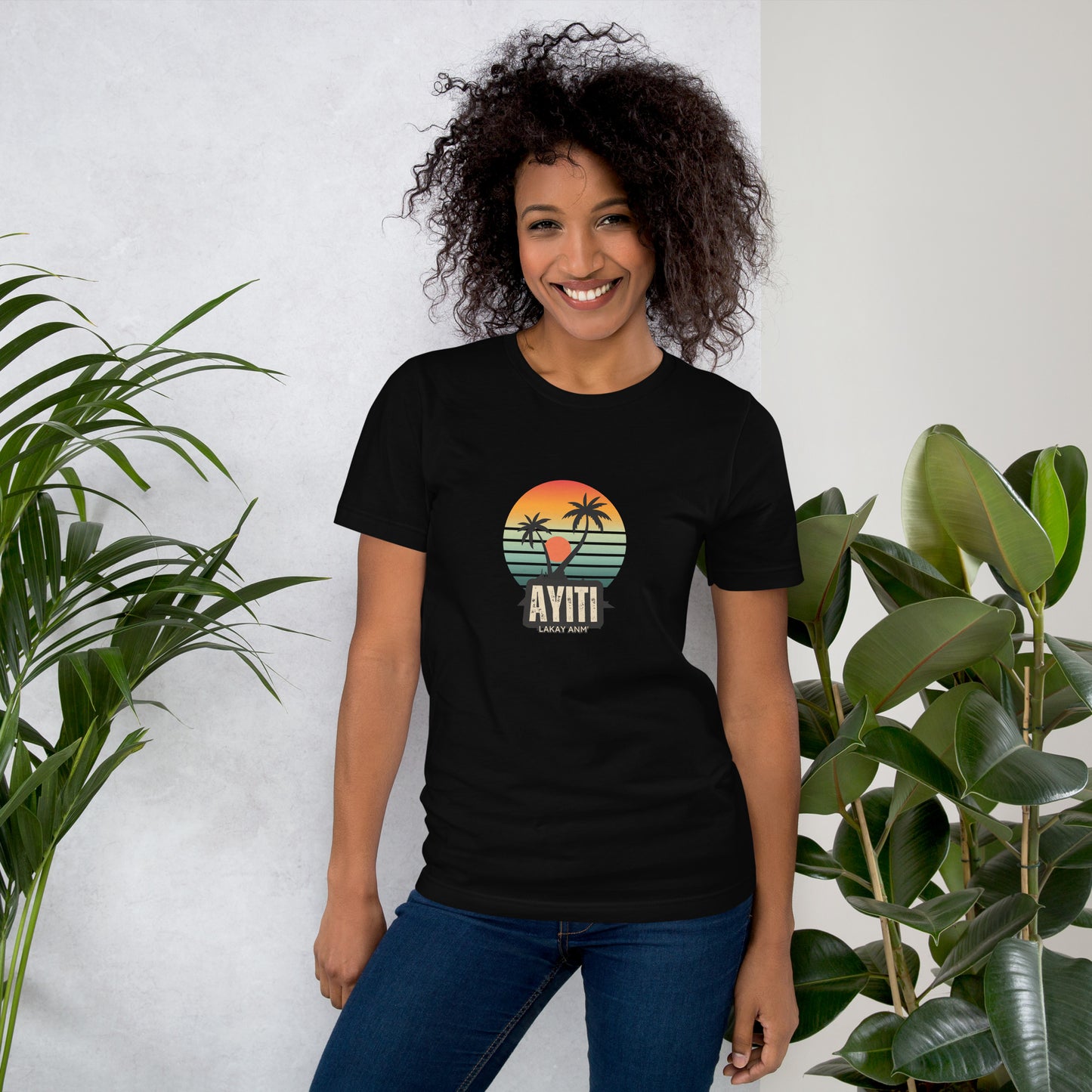 Smiling black woman wearing a black Bella+Canvas 'Ayiti Lakay Anm'' unisex T-shirt (front view) featuring a vibrant retro sunset graphic with two palm trees silhouetted. Green plants in the background.
