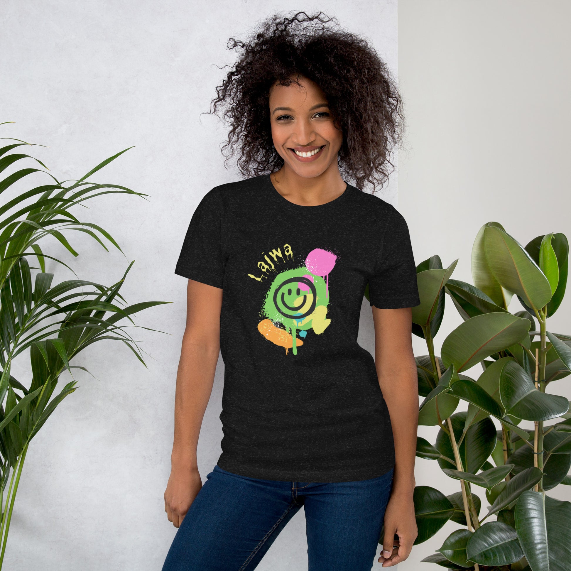 Smiling black female model wearing a black Bella+Canvas 'Lajwa' unisex T-shirt (front view) with a colorful dripping paint smiley face graphic in bright pink, green, orange, and yellow. She is standing in a room with green plants in the background.