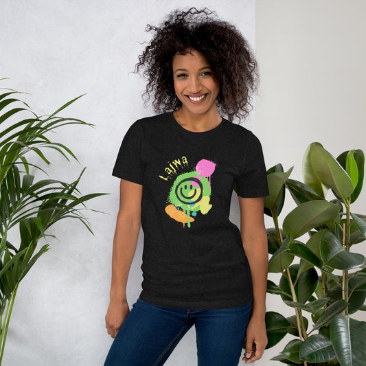 Smiling black female model wearing a black Bella+Canvas 'Lajwa' unisex T-shirt (front view) with a colorful dripping paint smiley face graphic in bright pink, green, orange, and yellow. She is standing in a room with green plants in the background.