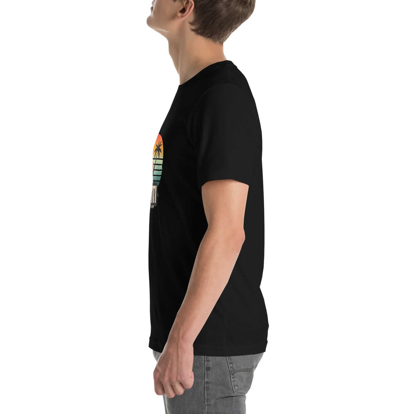 White man wearing a black Bella+Canvas 'Ayiti Lakay Anm'' unisex T-shirt (left side view) featuring a vibrant retro sunset graphic with two palm trees silhouetted. White background.