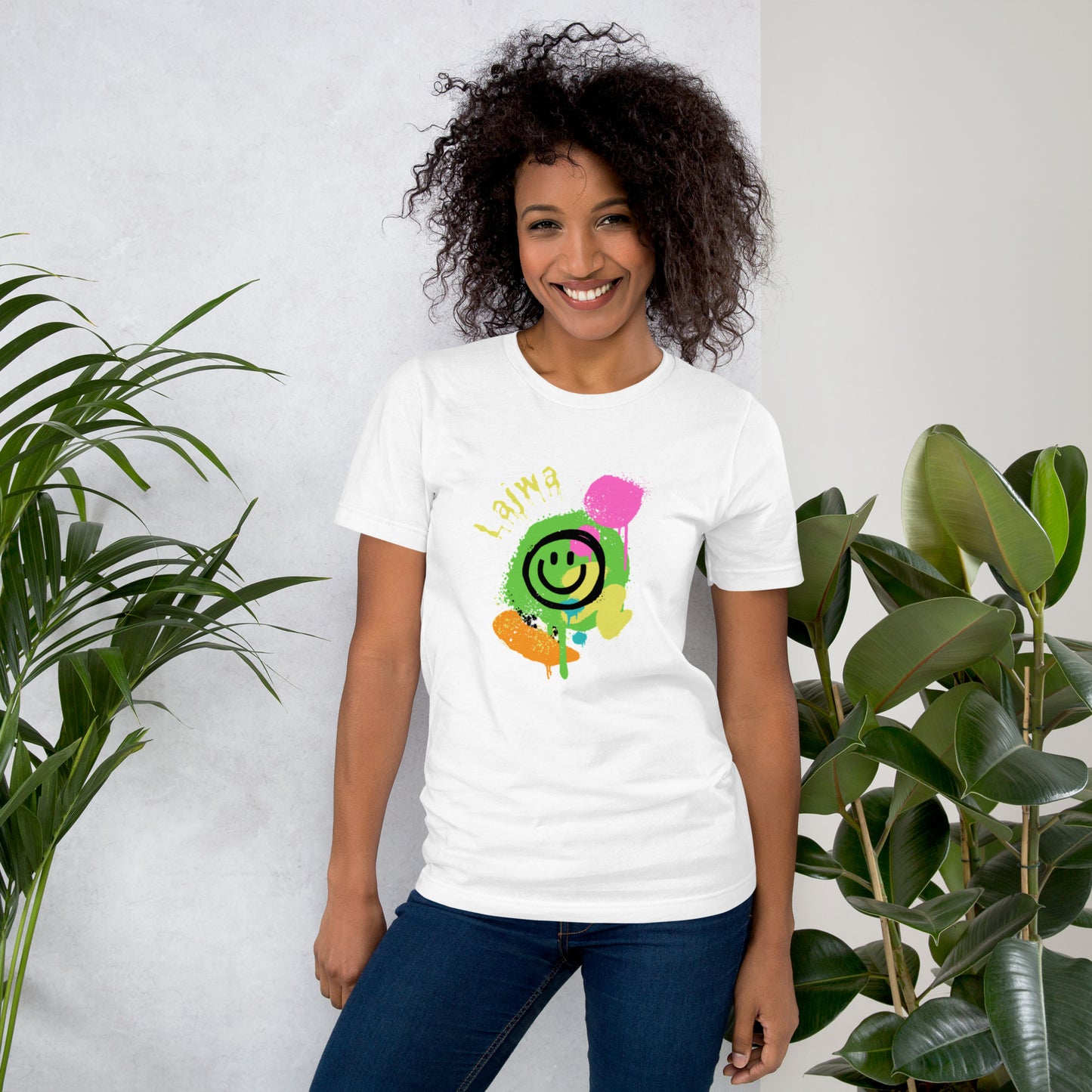 Smiling black female model wearing a white Bella+Canvas 'Lajwa' unisex T-shirt (front view) with a colorful dripping paint smiley face graphic in bright pink, green, orange, and yellow. She's standing in a room with green plants in the background.