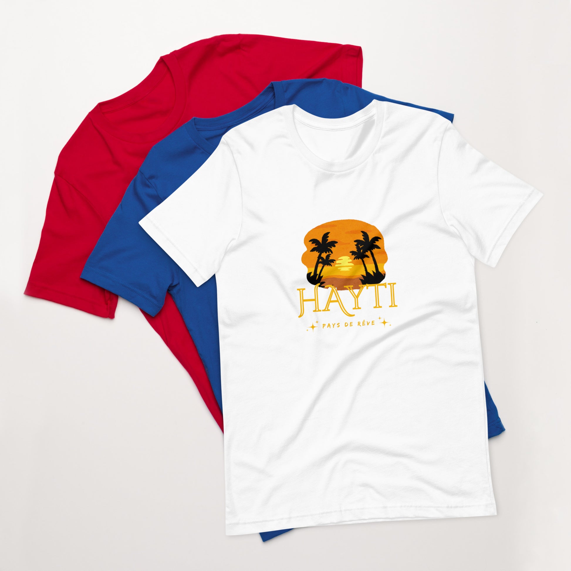 A white Bella+Canvas 'HAYTI PAYS DE RÊVE' unisex T-shirt (front view) featuring a golden hour sunset graphic with palm trees and twinkling stars. Red and bleu T-shirt in the background.