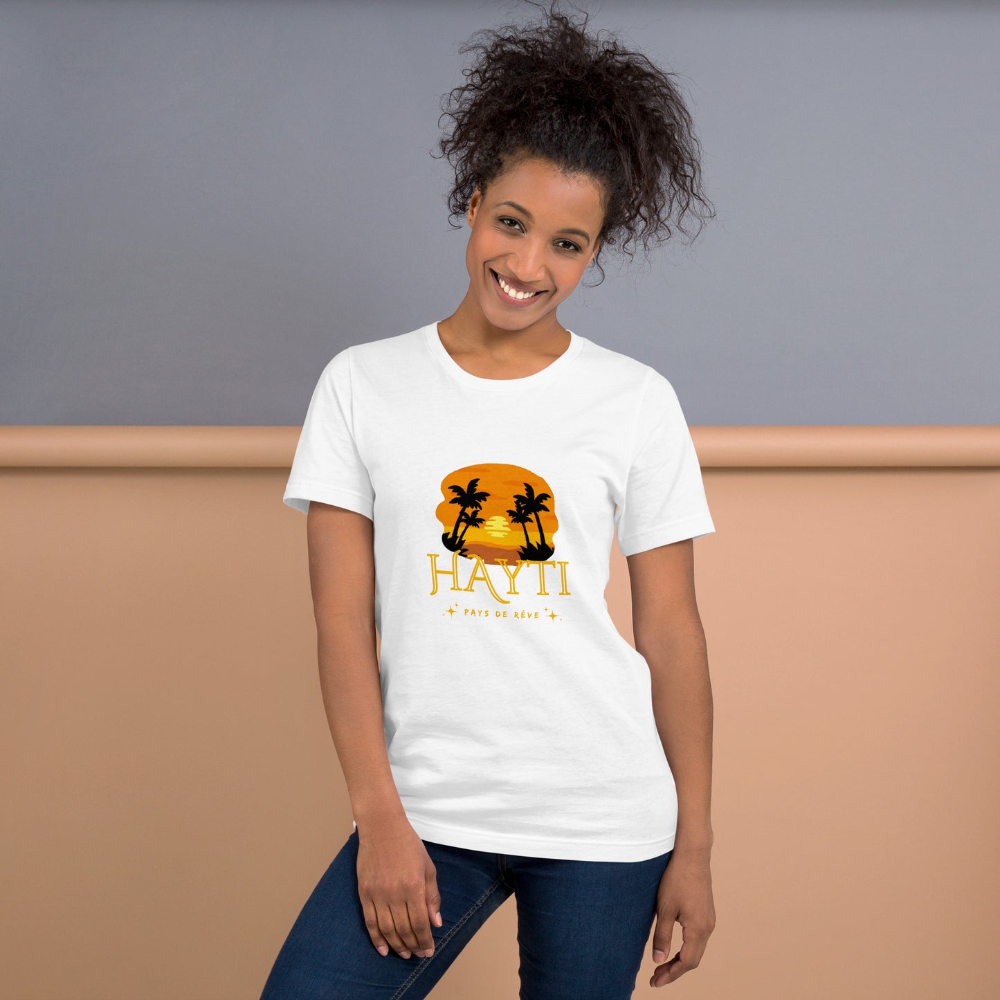 Smiling woman wearing a white Bella+Canvas 'HAYTI PAYS DE RÊVE' unisex T-shirt (front view) featuring a golden hour sunset graphic with palm trees and twinkling stars, in a room with Grey and beige background.