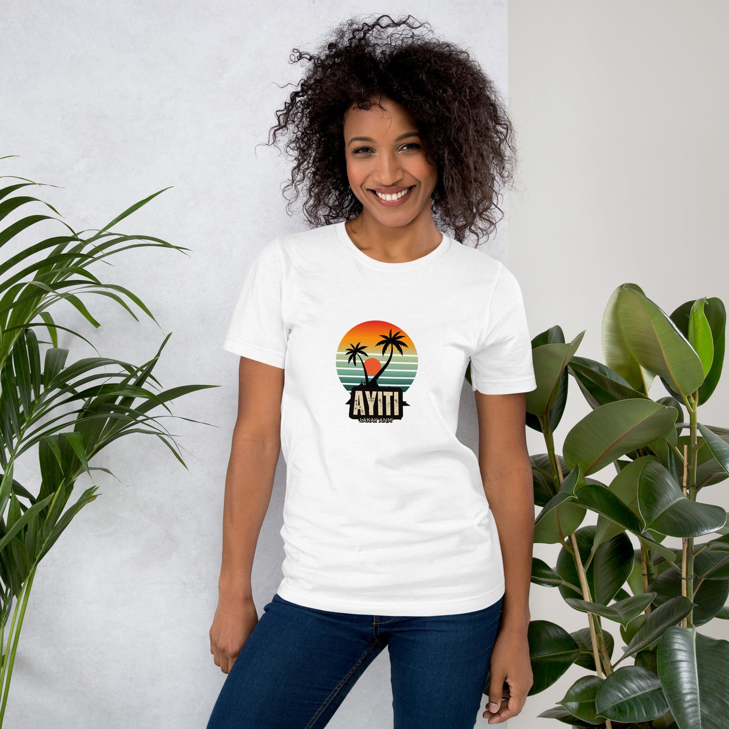 Smiling woman wearing a white Bella+Canvas 'Ayiti Lakay Anm' unisex T-shirt (front view) featuring a vibrant retro sunset graphic with two palm trees silhouetted. With green plants in the background.