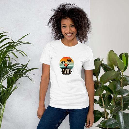 Smiling woman wearing a white Bella+Canvas 'Ayiti Lakay Anm' unisex T-shirt (front view) featuring a vibrant retro sunset graphic with two palm trees silhouetted. With green plants in the background.