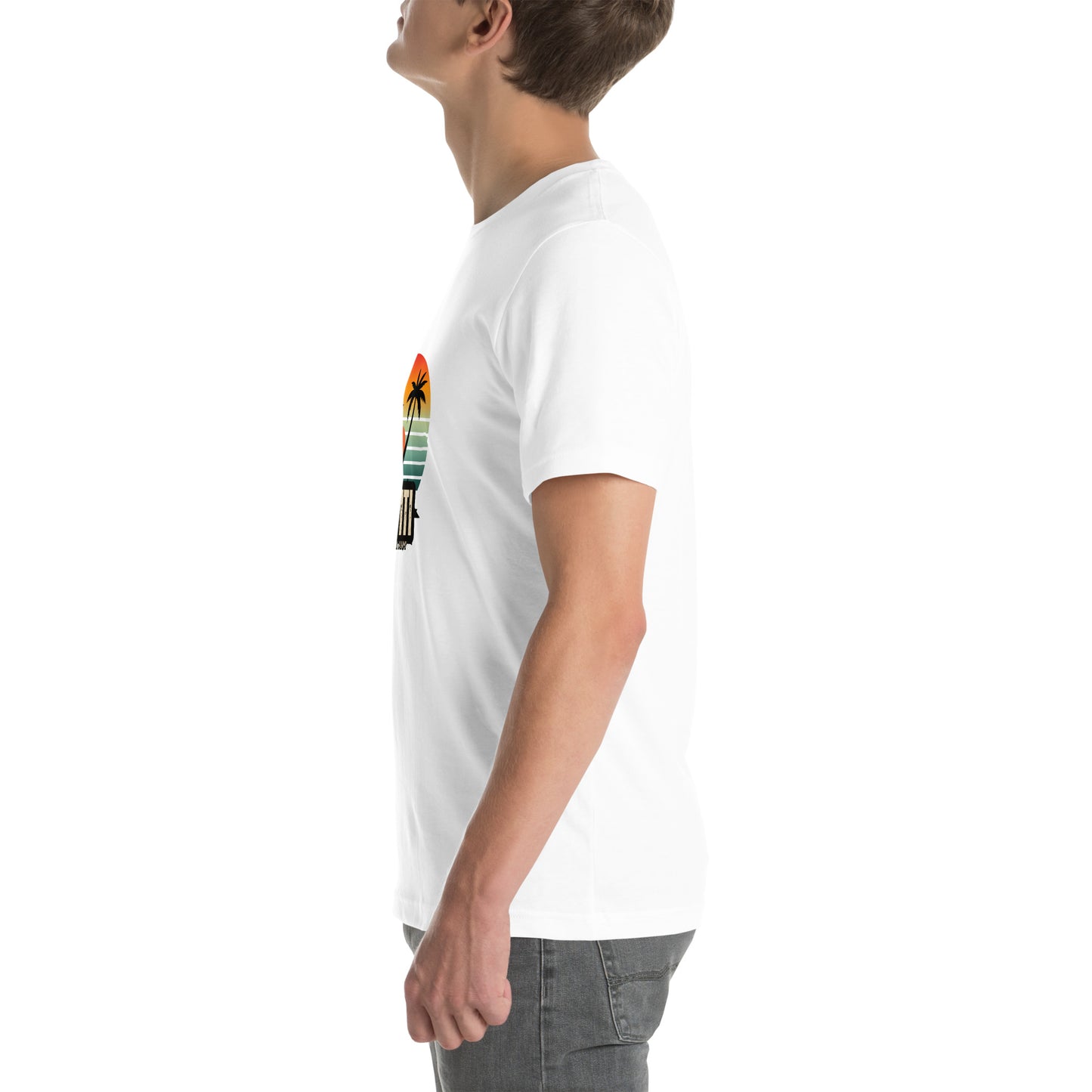White man wearing a white Bella+Canvas 'Ayiti Lakay Anm'' unisex T-shirt (left side view) featuring a vibrant retro sunset graphic with two palm trees silhouetted.