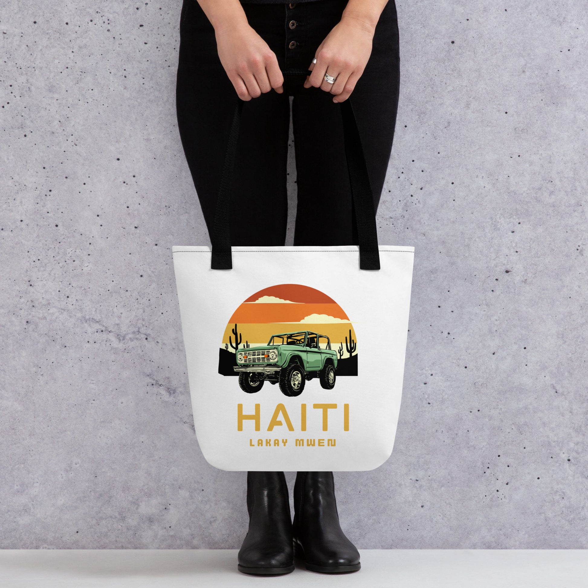 Model hands holding a white canvas tote bag with black handles, featuring a circular orange and yellow sunset graphic with a green vintage Jeep, black mountains, and cactus silhouettes. The text 'HAITI' is below the graphic, with 'LAKAY MWEN' underneath it.