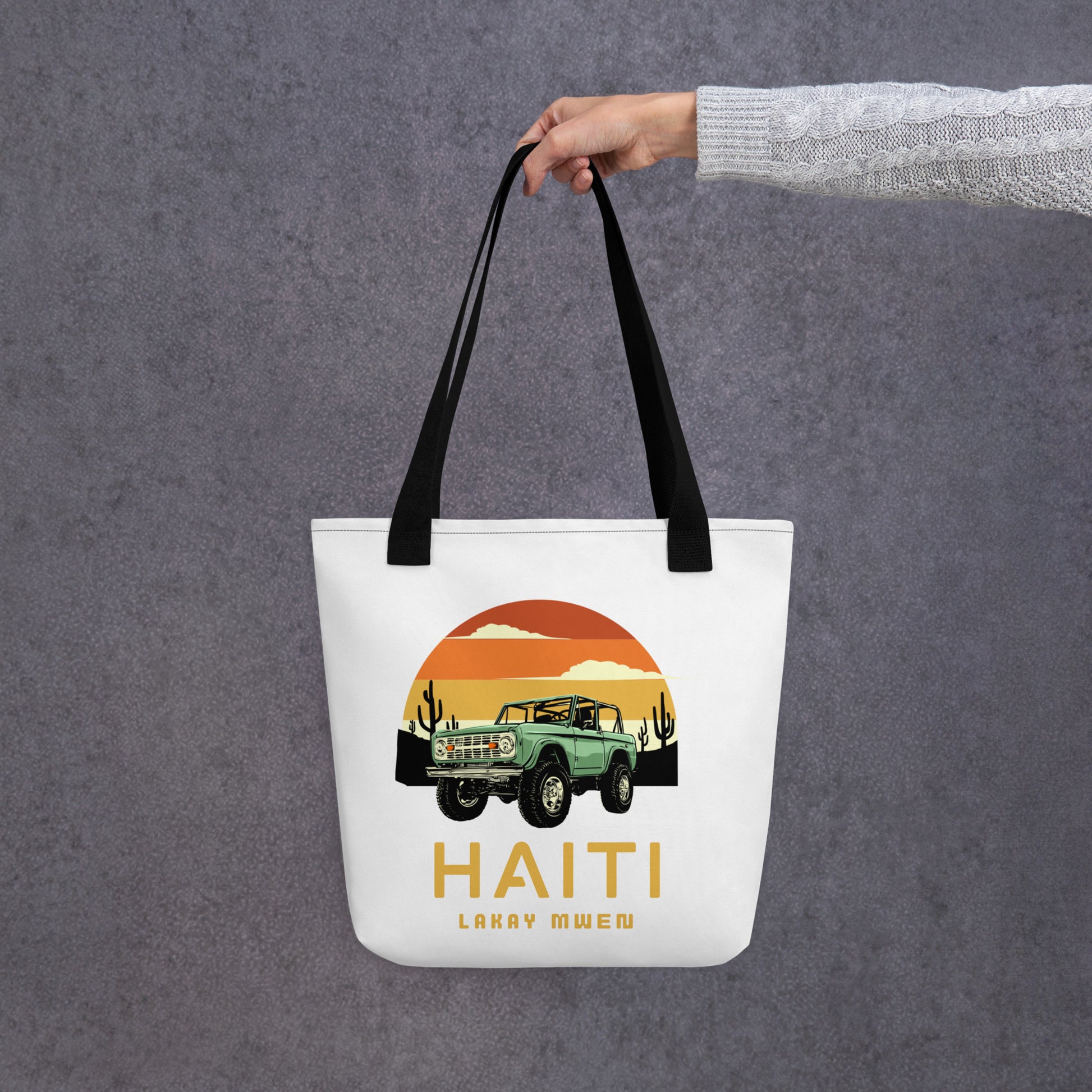 White canvas tote bag with black handles, featuring a circular orange and yellow sunset graphic with a green vintage Jeep, black mountains, and cactus silhouettes. The text 'HAITI' is below the graphic, with 'LAKAY MWEN' underneath it.