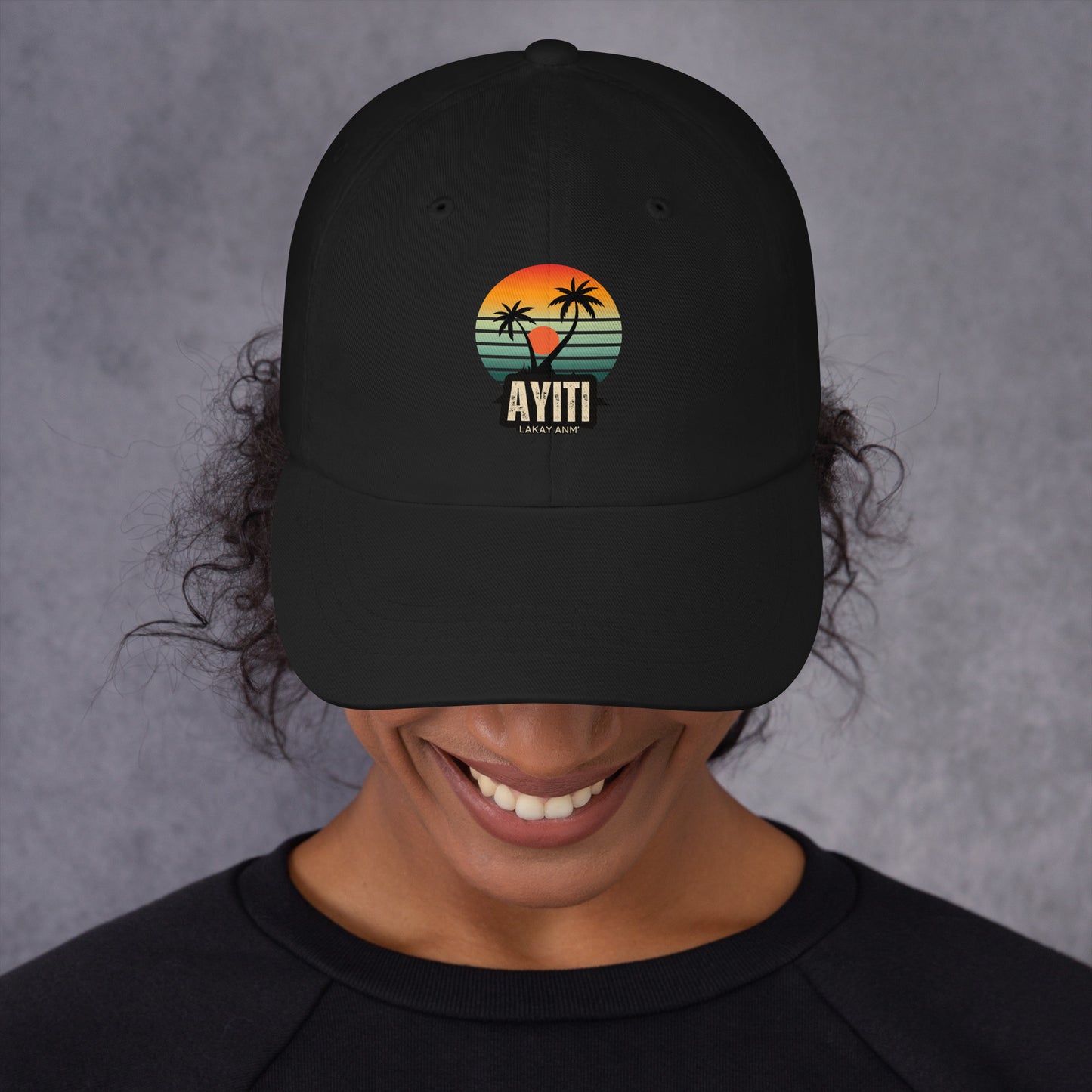 Close-up of a person wearing a black baseball cap with a circular graphic on the front featuring an orange and yellow sunset over blue and green stripes, silhouetted by two palm trees. The text 'AYITI' and 'LAKAY ANM'' is below the graphic in a stylized font. The person is smiling slightly.