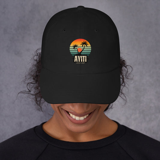Close-up of a person wearing a black baseball cap with a circular graphic on the front featuring an orange and yellow sunset over blue and green stripes, silhouetted by two palm trees. The text 'AYITI' and 'LAKAY ANM'' is below the graphic in a stylized font. The person is smiling slightly.