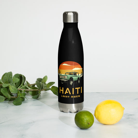 A black insulated water bottle with a silver cap and base, featuring a circular orange and yellow sunset graphic with a green vintage Jeep, black mountains, and cactus silhouettes. The text 'HAITI' is below the graphic in orange, with 'LAKAY MWEN' underneath it. A lime and a lemon with a bunch of mint next to it.