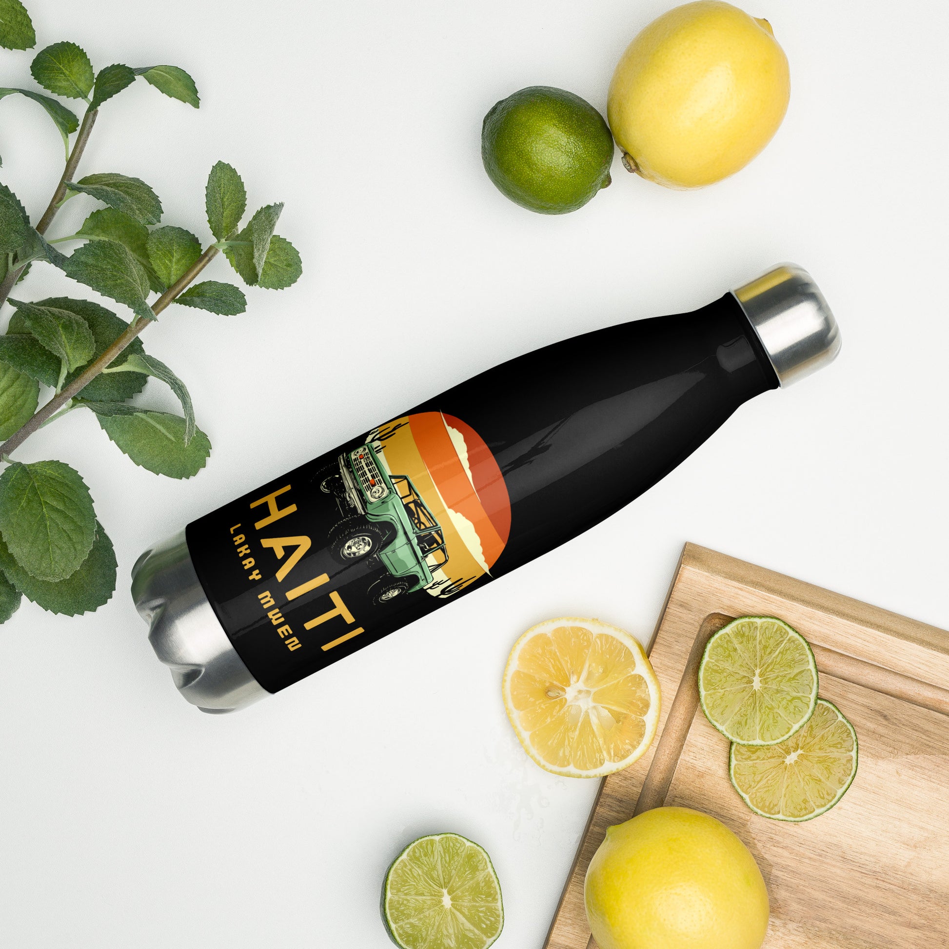 A black insulated water bottle with a silver cap and base, featuring a circular orange and yellow sunset graphic with a green vintage Jeep, black mountains, and cactus silhouettes. The text 'HAITI' is below the graphic in orange, with 'LAKAY MWEN' underneath it.. A mint bunch, a lime and lime slices, and two lemons and a lemon slice are on the surface next to the bottle.