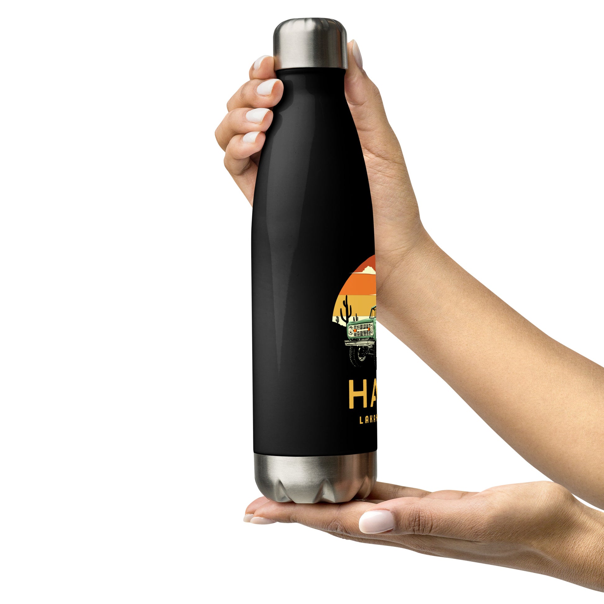 A black insulated water bottle with a silver cap and base, featuring a circular orange and yellow sunset graphic with a green vintage Jeep, black mountains, and cactus silhouettes. The text 'HAITI' is below the graphic in orange, with 'LAKAY MWEN' underneath it. Two hands are showing holding the bottle, slightly turned, showing its right side