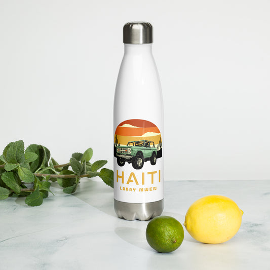 A white insulated water bottle with a silver cap and base, featuring a circular orange and yellow sunset graphic with a green vintage Jeep, black mountains, and cactus silhouettes. The text 'HAITI' is below the graphic in orange, with 'LAKAY MWEN' underneath it. Mint bunch, a lime, and a lemon are on the surface next to the bottle.