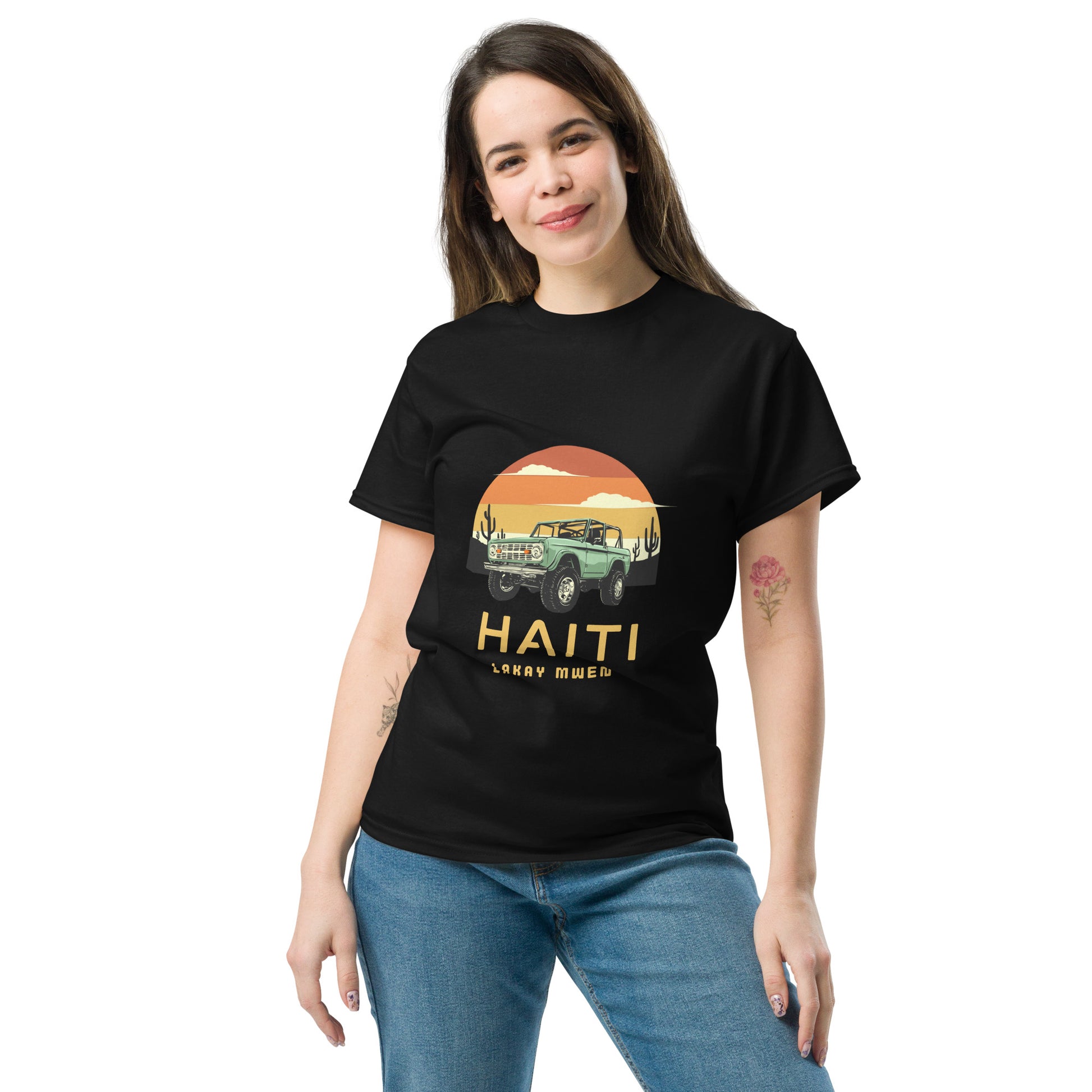 Smiling White women standing wearing a black Bella+Canvas 'Haiti Lakay Mwen' unisex T-shirt featuring a retro sunset and green vintage Jeep graphic. White background. 