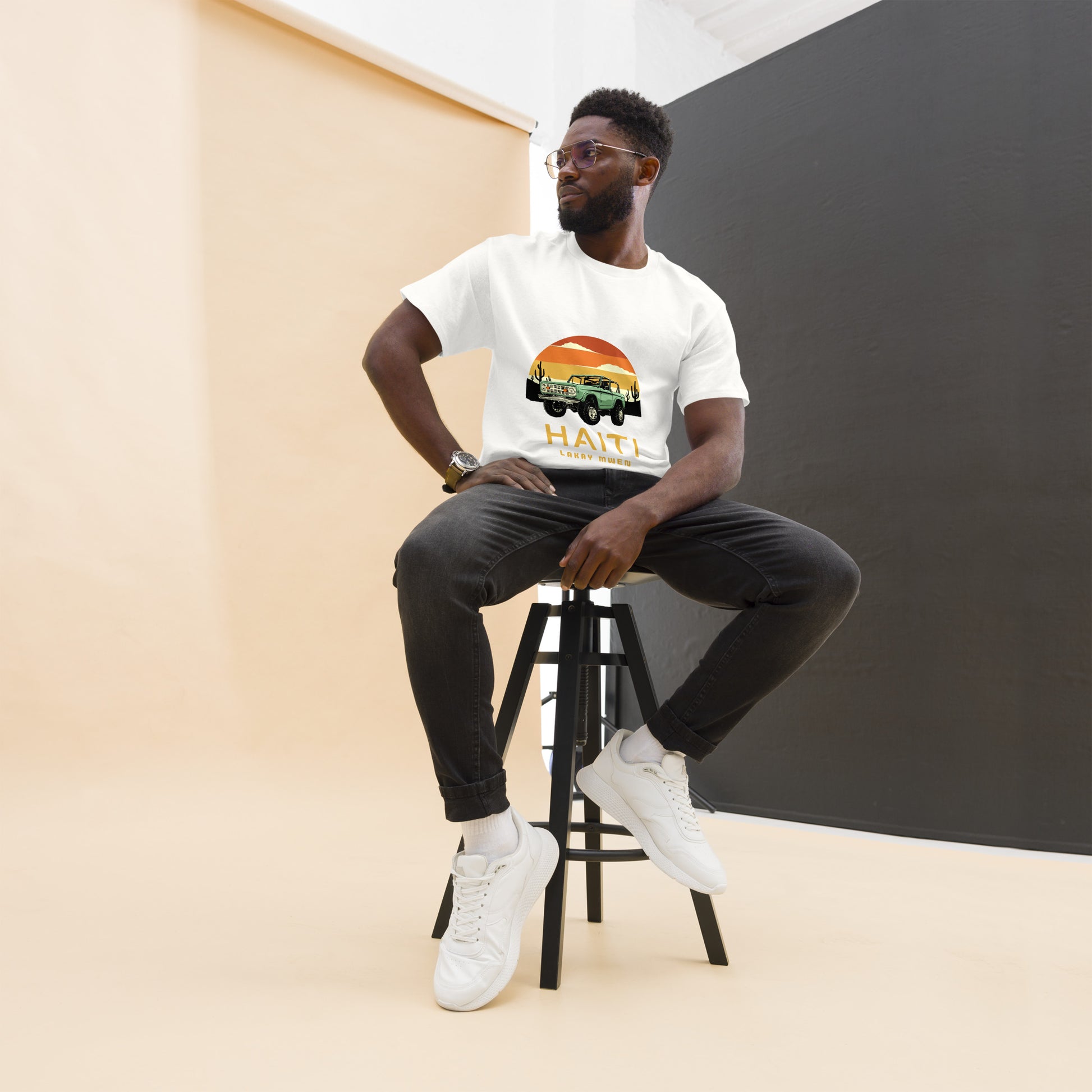 Male model sitting on a stool in a studio room wearing Bella+Canvas 'Haiti Lakay Mwen' unisex T-shirt featuring a retro sunset and green vintage Jeep graphic. Gray and beige background. 