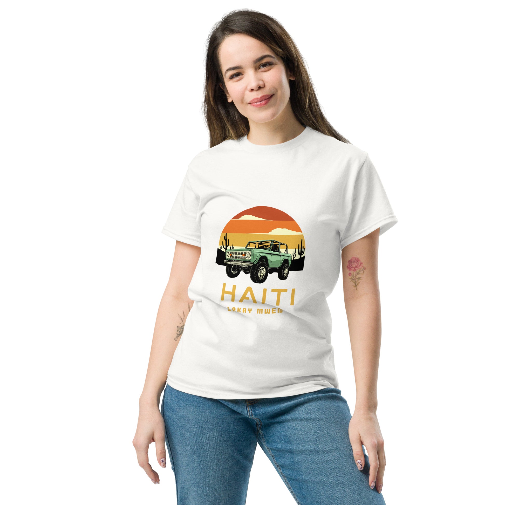 White woman standing and smiling wearing a Bella+Canvas 'Haiti Lakay Mwen' unisex T-shirt featuring a retro sunset and green vintage Jeep graphic. White background. 