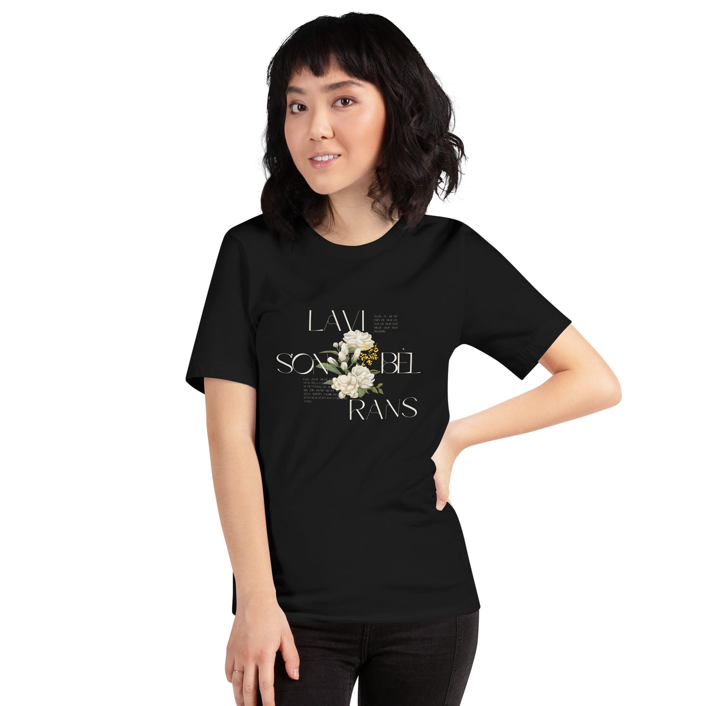 Female model wearing a black Bella+Canvas 'LAVI S'ON BÈL RANS' unisex T-shirt (front view) featuring cream text and a realistic white floral graphic. The man has a bob cut with bangs and is wearing black jeans.