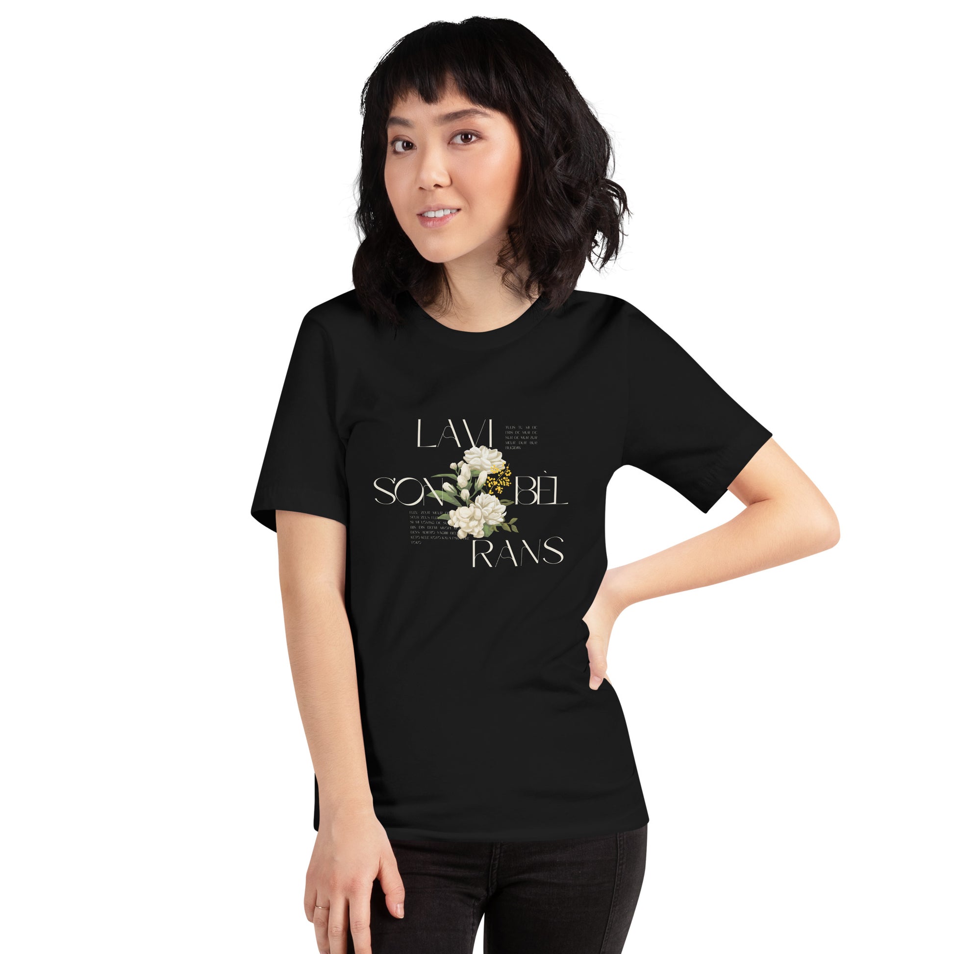 Female model wearing a black Bella+Canvas 'LAVI S'ON BÈL RANS' unisex T-shirt (front view) featuring cream text and a realistic white floral graphic. The man has a bob cut with bangs and is wearing black jeans.