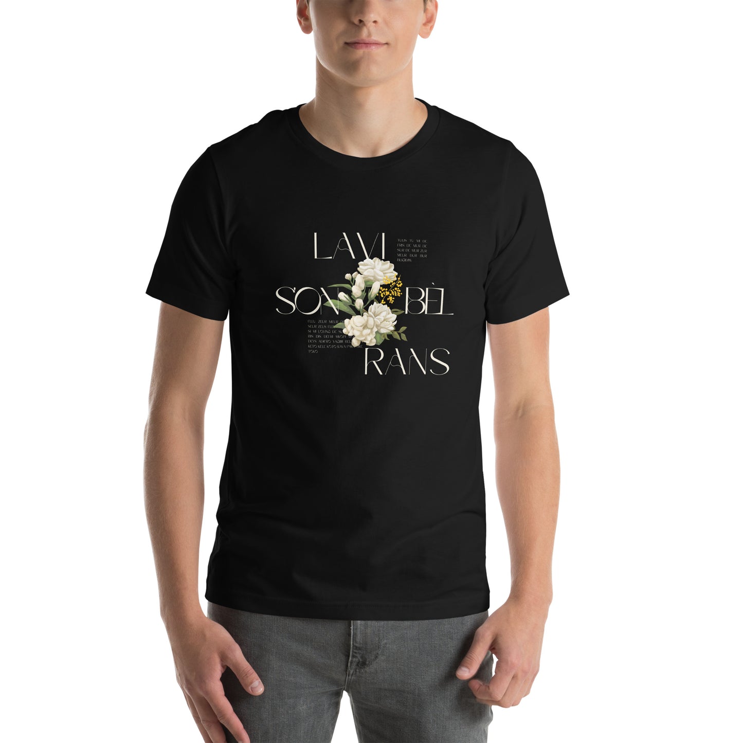 Male model wearing a black Bella+Canvas 'LAVI S'ON BÈL RANS' unisex T-shirt (front view) featuring cream text and a realistic white floral graphic. Wearing Grey jeans. White background. 