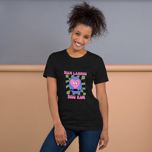 Woman wearing a black Bella+Canvas 'Nan Lanmou Nou Kwe' unisex T-shirt (front view) featuring a pink and blue abstract heart graphic with a smiley face and paint drip accents. She has short, curly hair and is smiling.