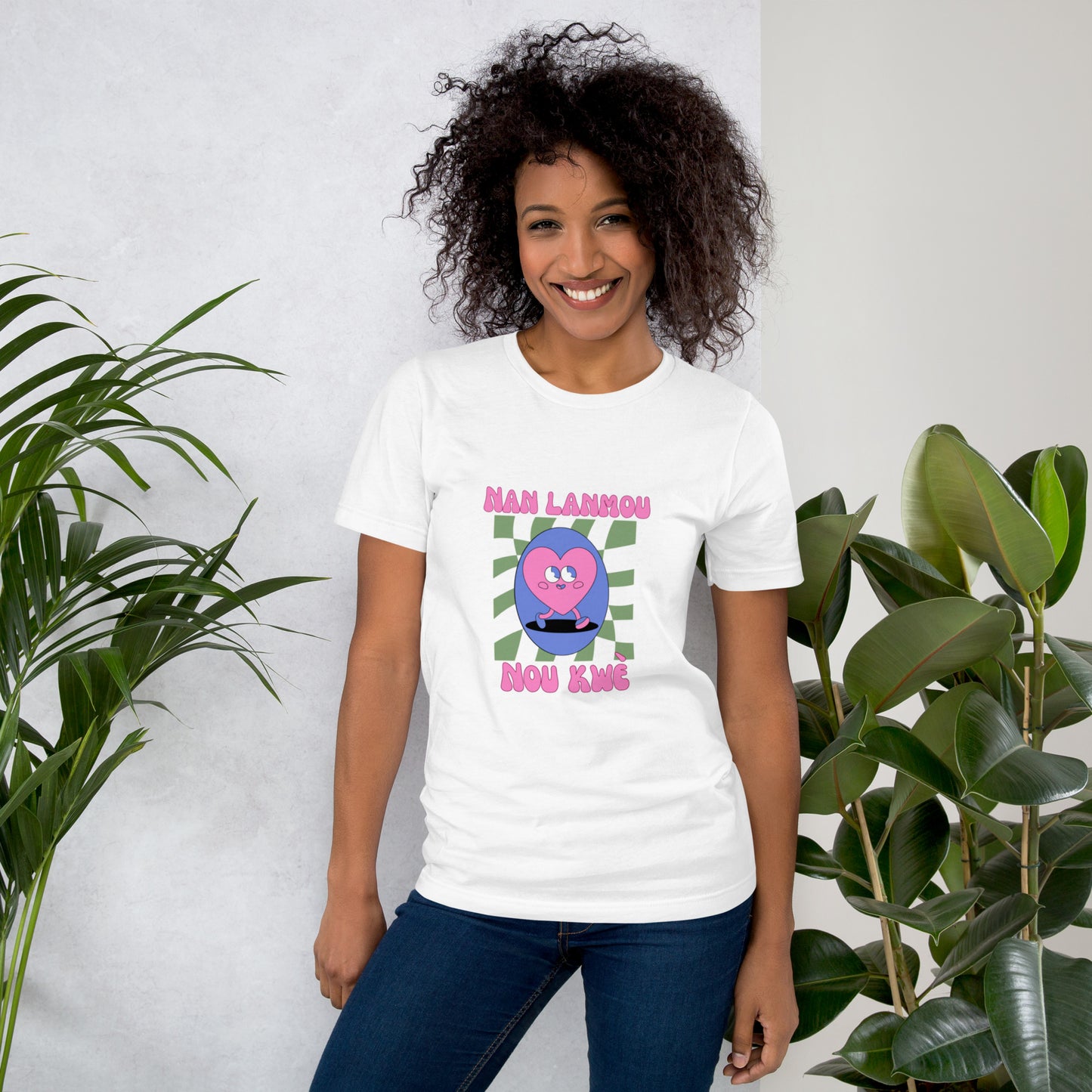 Woman wearing a white Bella+Canvas 'Nan Lanmou Nou Kwe' unisex T-shirt (front view) featuring a pink and blue abstract heart graphic with a smiley face and paint drip accents. She has short, curly hair and is smiling.