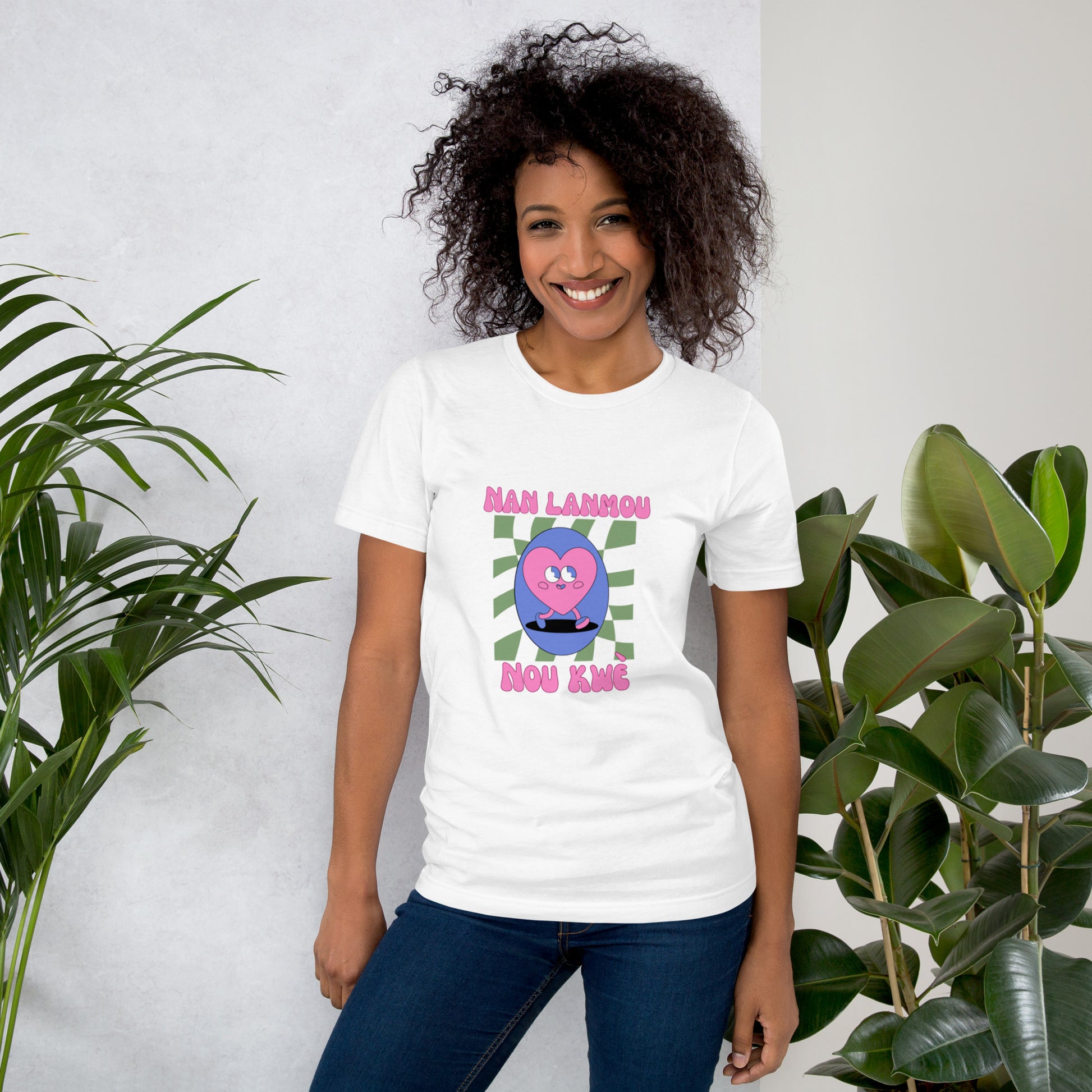 Woman wearing a white Bella+Canvas 'Nan Lanmou Nou Kwe' unisex T-shirt (front view) featuring a pink and blue abstract heart graphic with a smiley face and paint drip accents. She has short, curly hair and is smiling.